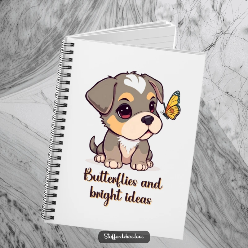 Funny Staffordshire pup with one ear flopped over, looking curiously at a fluttering butterfly on the cover of a lined notebook.